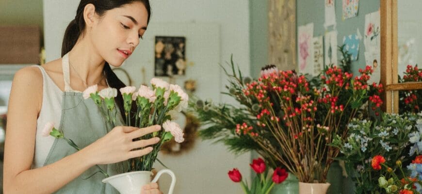 A woman holding a vase full of flowers in front of a shelf with multiple vases of flowers on it